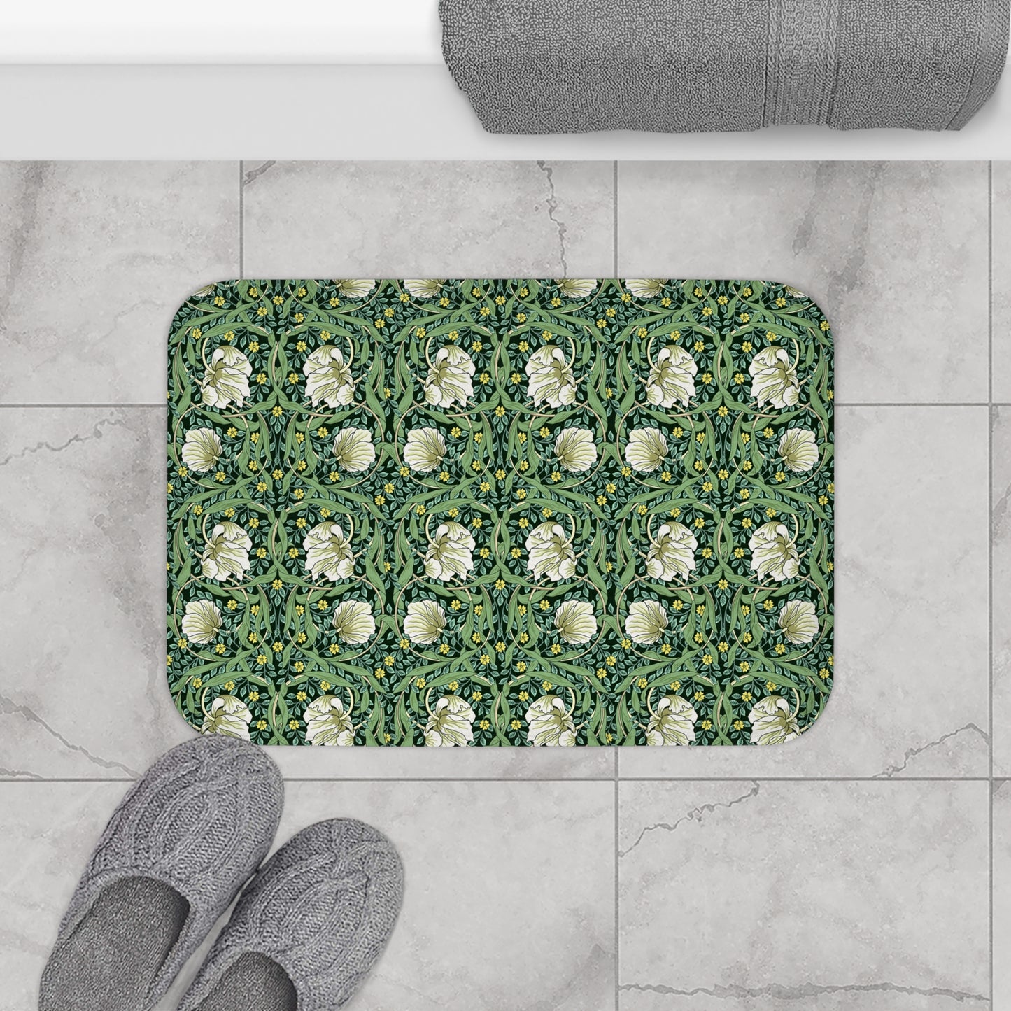 A rectangular bath mat with a green and white floral pattern is placed on a light gray marble tile floor next to gray slippers, with a folded gray towel partially visible at the top.
