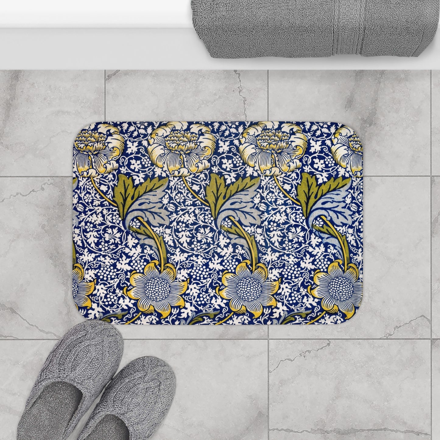Decorative bath mat with floral pattern on a tiled floor, next to slippers and a towel.