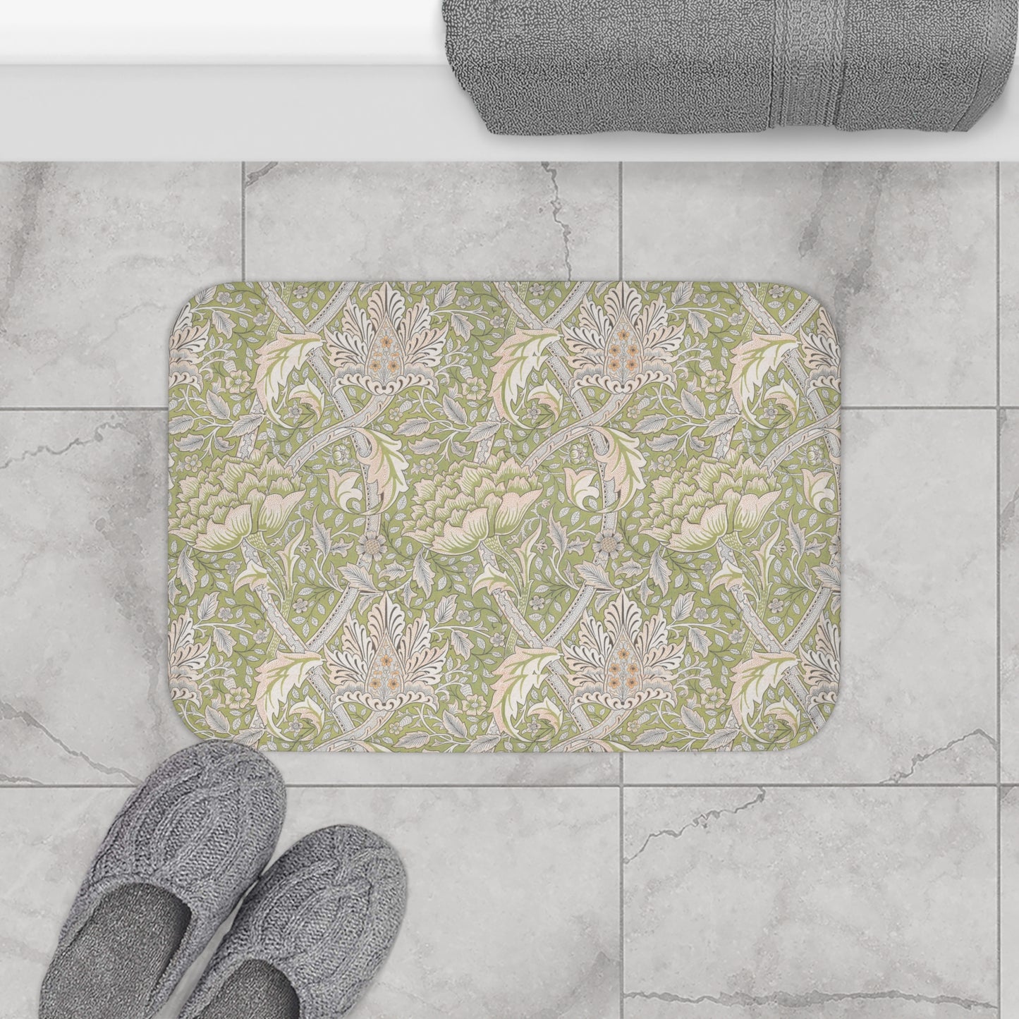 Decorative bath mat with floral pattern on a tiled floor, next to slippers and a towel.