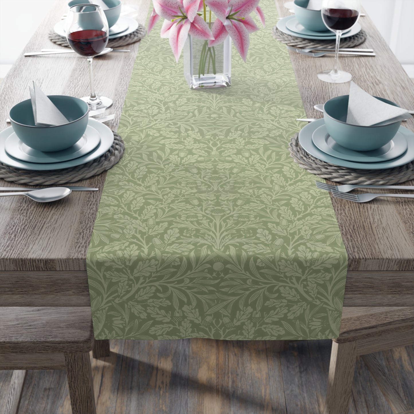 A green table runner with a floral William Morris & Co pattern, featuring acorns and oak leaves, laid out on a wooden dining table with cutlery and a vase of pink flowers in the background.