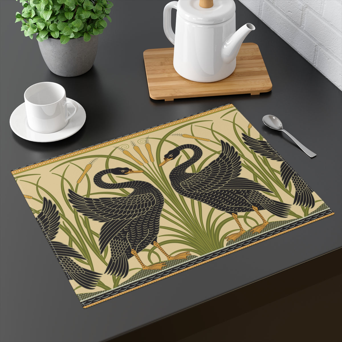 A rectangular table mat with a vintage-style illustration of two black swans among green reeds, placed on a dark tabletop next to a white teapot on a wooden board, a cup and saucer, and a spoon.