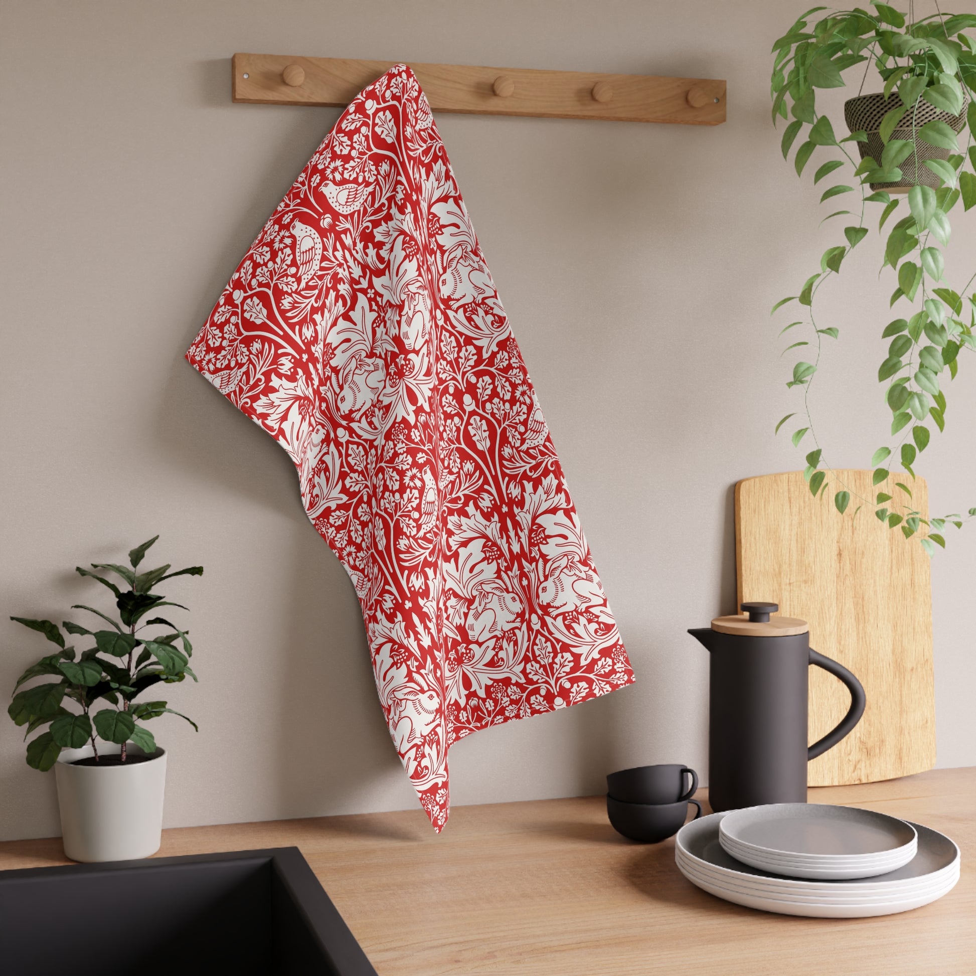 A red and white patterned kitchen towel hangs from a wooden peg on a beige wall above a kitchen counter with dishes, a black kettle, and potted plants.