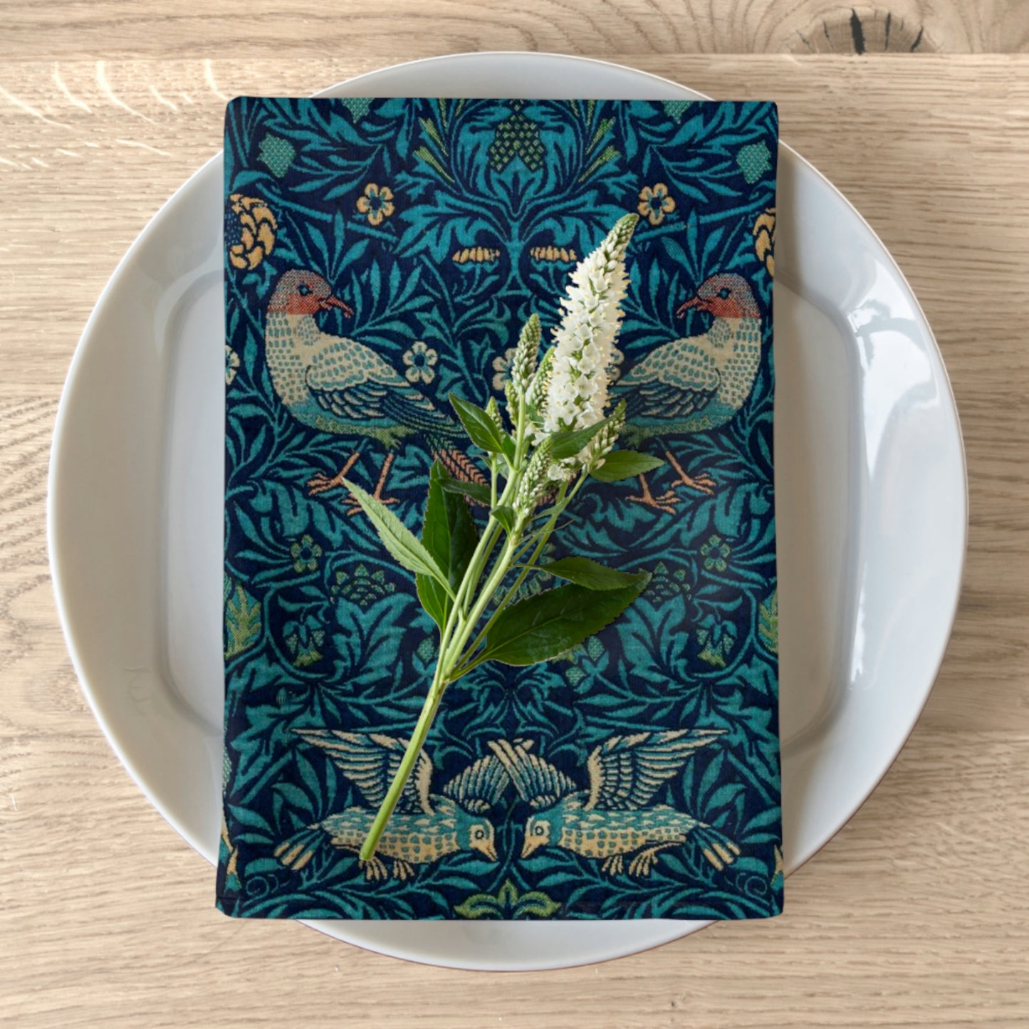 A blue cotton twill napkin with a floral and bird pattern is placed on a white plate on a wooden table, accompanied by a sprig of flowers.