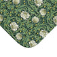 A close-up of a textile or wallpaper pattern featuring large white flowers, green leaves, and small yellow blossoms on a dark green background, with one corner folded over to show the material edge against a white studio background.