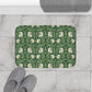 A rectangular bath mat with a green and white floral pattern is placed on a light gray marble tile floor next to gray slippers, with a folded gray towel partially visible at the top.