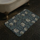 A rectangular bath mat with a William Morris-style floral pattern in muted blue and white, placed on dark tiled bathroom floor next to a white clawfoot tub.