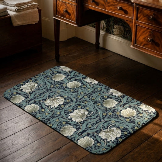 A rectangular area rug with a William Morris-style floral pattern in teal, navy, and cream, shown on a dark wood floor in a room with a wooden writing desk.
