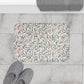 Floral-patterned bath mat on a tiled floor with slippers and a towel.