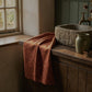 A rust-orange textured towel with a dark botanical leaf pattern draped over a rustic wooden bathroom counter next to a stone sink and a green ceramic vase.