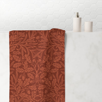 A terracotta-colored bath towel with a matching botanical damask pattern draped over a white marble sink, with a white tiled wall and minimalist bathroom accessories in the background.