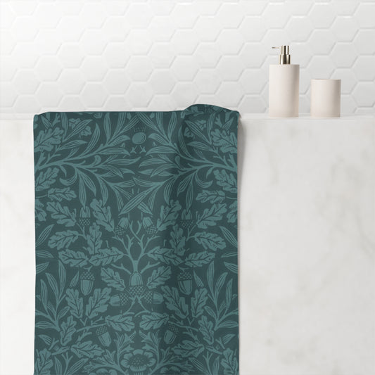 A dark teal bathroom towel with a classic William Morris-style floral and botanical damask pattern, draped over a white marble counter next to minimalist white ceramic bathroom accessories.