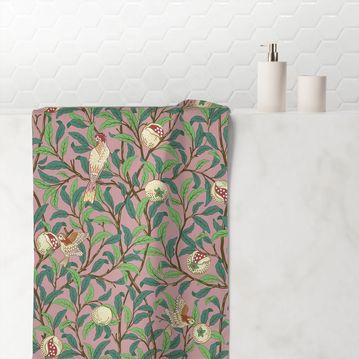 A bath towel with a William Morris-style botanical pattern featuring green leaves, brown branches, and birds on a dusty pink background, draped over a white marble counter next to a white tiled wall.