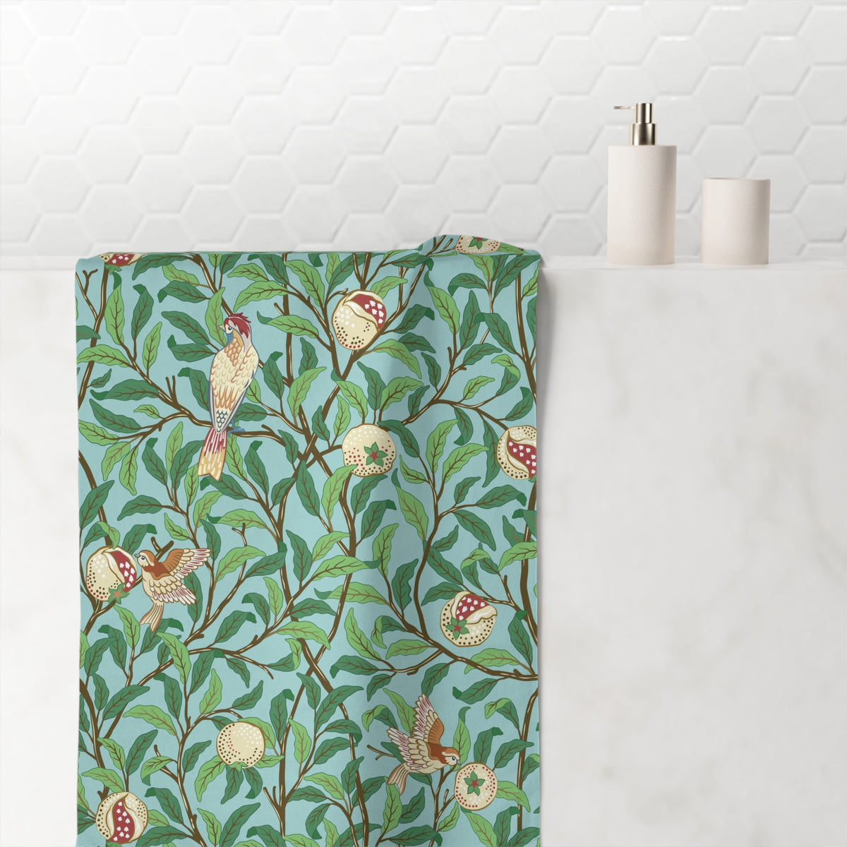 A folded bath towel with a William Morris-style botanical and bird pattern in green, brown, cream, and red, draped over a white marble counter next to a white tiled wall and bathroom accessories.