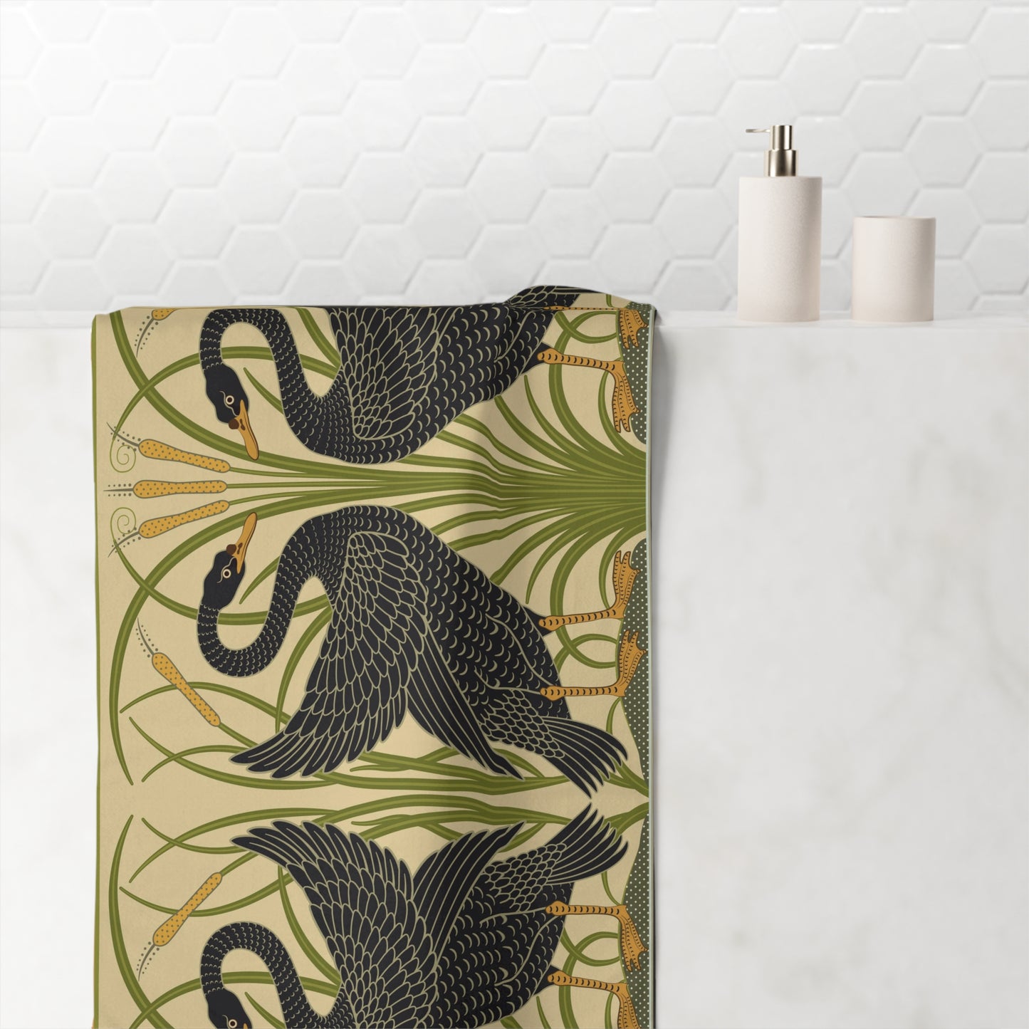 A folded bath towel with a repeating pattern of black cranes and green reeds on a beige background, styled on a white marble countertop next to a soap dispenser and cup against a white hexagonal tile backsplash.