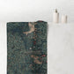A rectangular bath towel with a detailed William Morris-style woodland print featuring deer and foliage, draped over the edge of a white marble bathroom counter next to a white tiled wall.
