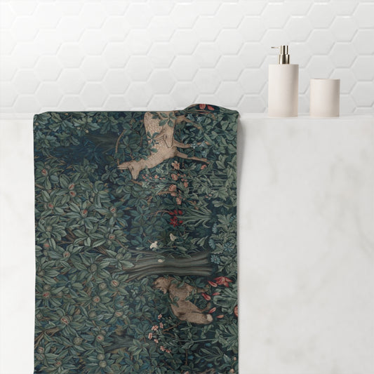A rectangular bath towel with a detailed William Morris-style woodland print featuring deer and foliage, draped over the edge of a white marble bathroom counter next to a white tiled wall.