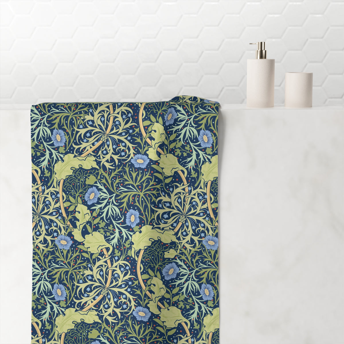 A folded bath towel with a William Morris-style floral and leaf pattern in navy blue, sage green, and light blue, draped over a white marble bathroom counter next to a soap dispenser and cup.