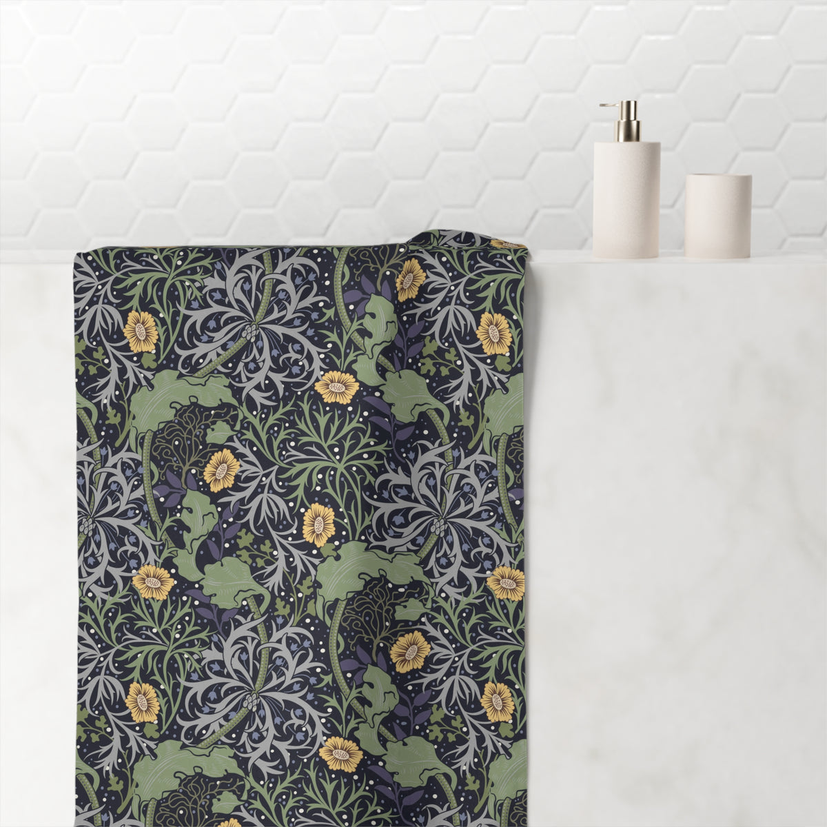 A bath towel with a detailed William Morris-style floral and scroll pattern in muted green, navy, gray, and yellow, draped over a white marble bathroom counter next to minimalist white ceramic bottles.