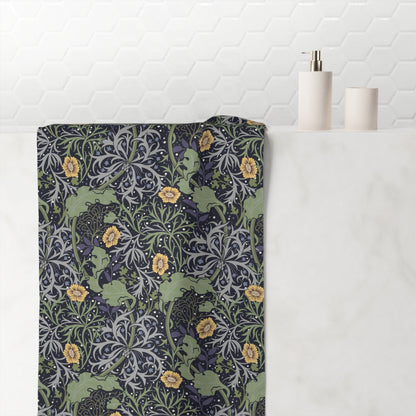 A bath towel with a detailed William Morris-style floral and scroll pattern in muted green, navy, gray, and yellow, draped over a white marble bathroom counter next to minimalist white ceramic bottles.