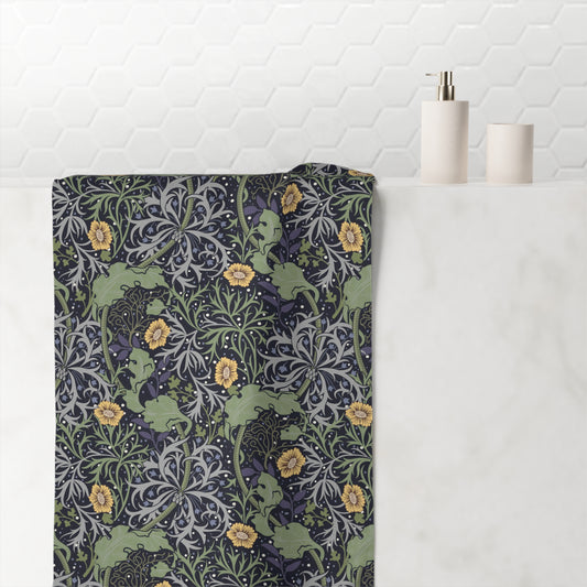 A bath towel with a detailed William Morris-style floral and scroll pattern in muted green, navy, gray, and yellow, draped over a white marble bathroom counter next to minimalist white ceramic bottles.