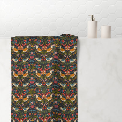 A bathroom towel draped over a white marble tub edge, featuring a dense floral and bird pattern on a dark background. The pattern includes orange birds, pink flowers, and green foliage.