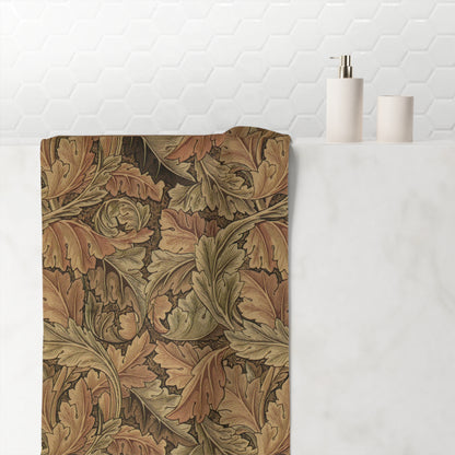 A rectangular bath mat with an intricate William Morris-style oak leaf pattern in earthy brown, beige, and muted green tones, styled on a white marble bathroom counter next to minimalist white toiletries against a white hexagonal tile wall.