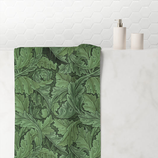 A bath mat featuring a classic green William Morris-style acanthus leaf pattern, styled on a white marble bathroom counter next to minimalist white ceramic bottles against a white hexagonal tile backsplash.