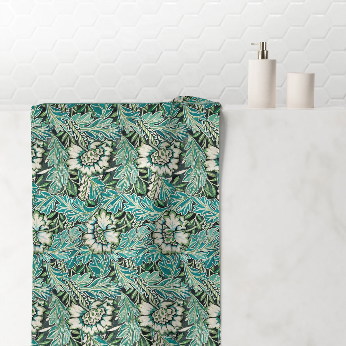 A bath towel with a detailed botanical pattern in teal, green, and white hangs over the edge of a white marble bathroom counter. In the background, a white hexagonal tiled wall is visible, along with a white pump dispenser and a small white cup.