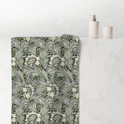 A bathroom towel with a detailed botanical pattern in shades of green, gray, and white, draped over a white marble counter next to minimalist white bathroom accessories.