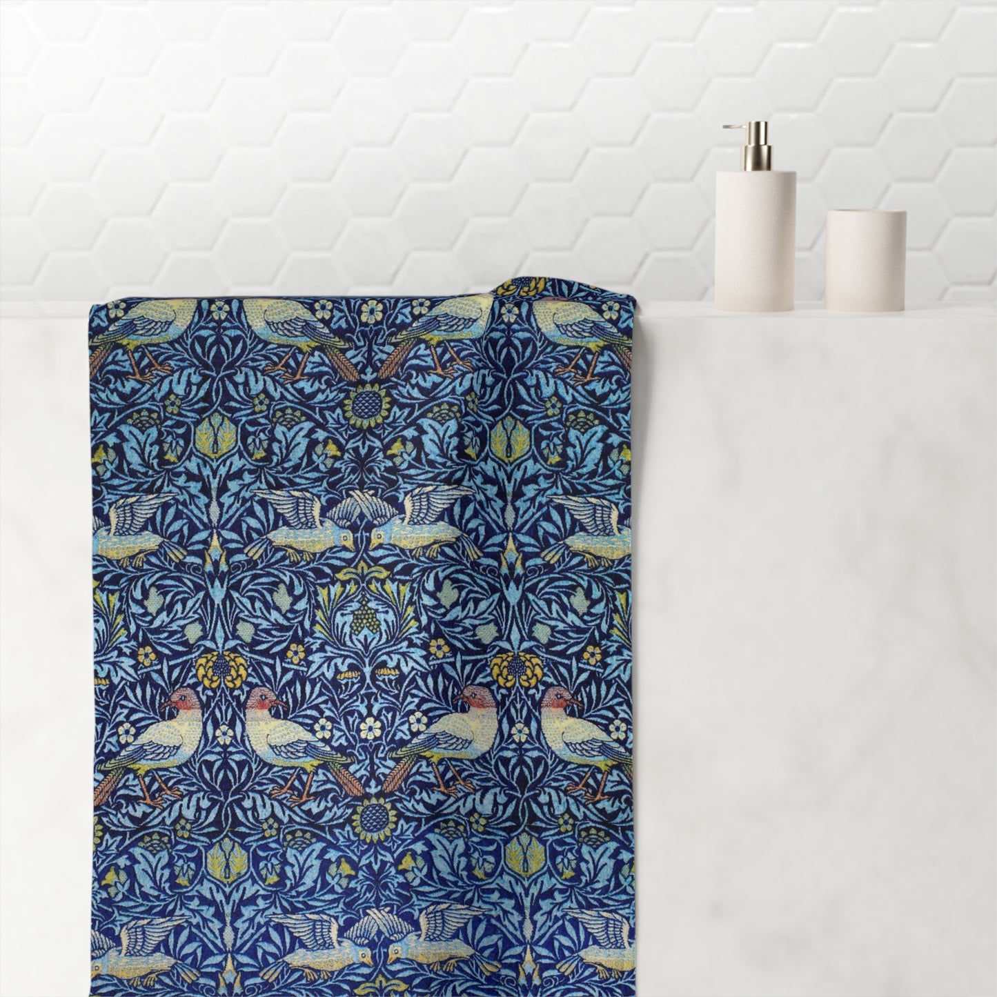 A bath towel with a detailed William Morris-style botanical and bird pattern in navy blue, light blue, yellow, and pink, draped over a white marble bathroom counter next to a white tiled wall.
