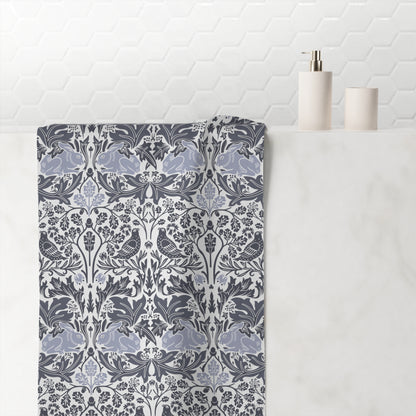 A folded bath towel with a detailed botanical damask pattern in slate blue, gray, and white, draped over a white marble bathroom counter next to minimalist white ceramic bottles.