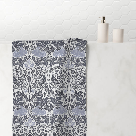 A folded bath towel with a detailed botanical damask pattern in slate blue, gray, and white, draped over a white marble bathroom counter next to minimalist white ceramic bottles.