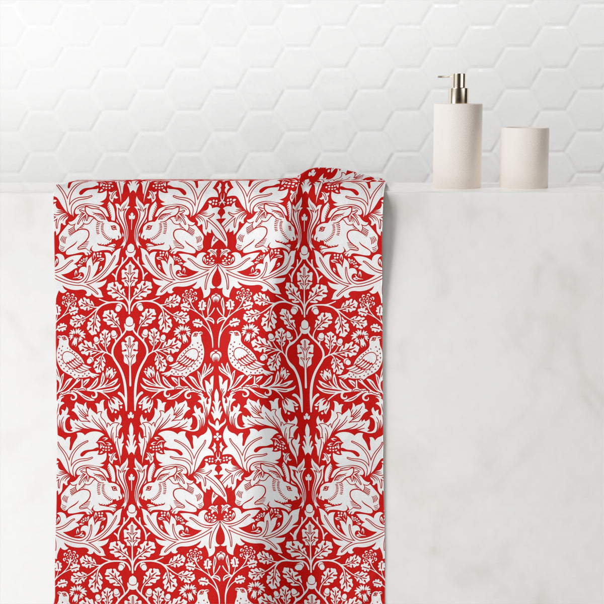 A red and white damask-patterned bath towel draped over a white marble bathroom counter, with a white hexagonal tile backsplash and minimalist soap dispenser in the background.