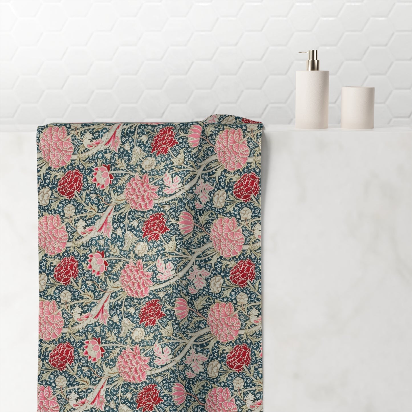A bath towel with a William Morris-style floral pattern in pink, red, and cream on a dark blue background, draped over a white marble bathroom counter next to a soap dispenser and cup.