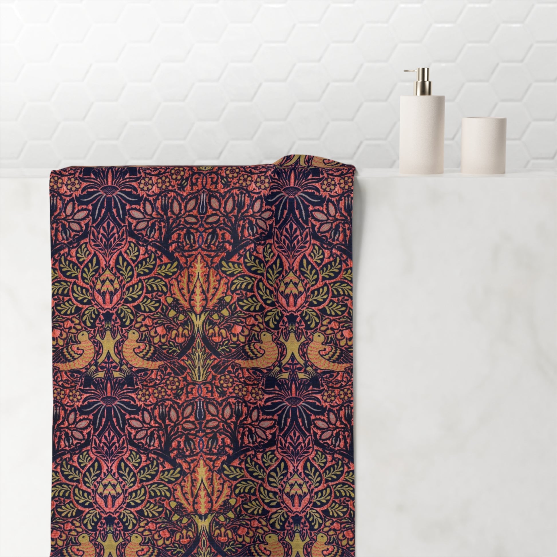 A folded bath towel with an intricate, symmetrical botanical and bird pattern in dark navy, coral, green, and gold, draped over a white marble bathroom counter next to a white soap dispenser.