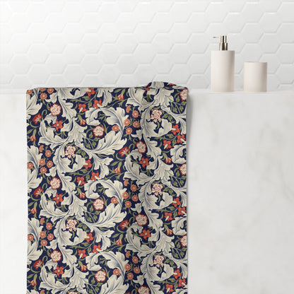 A folded bath towel with a William Morris-style floral and scroll pattern in navy blue, cream, coral, and green, draped over the edge of a white marble bathroom counter next to a soap dispenser and cup.