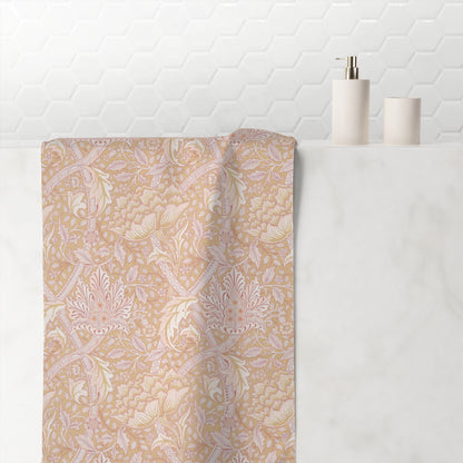 A beige bath towel with a delicate pink and white botanical damask pattern draped over a white marble ledge in a bathroom setting, with a white hexagonal tile wall and minimalist soap dispensers in the background.