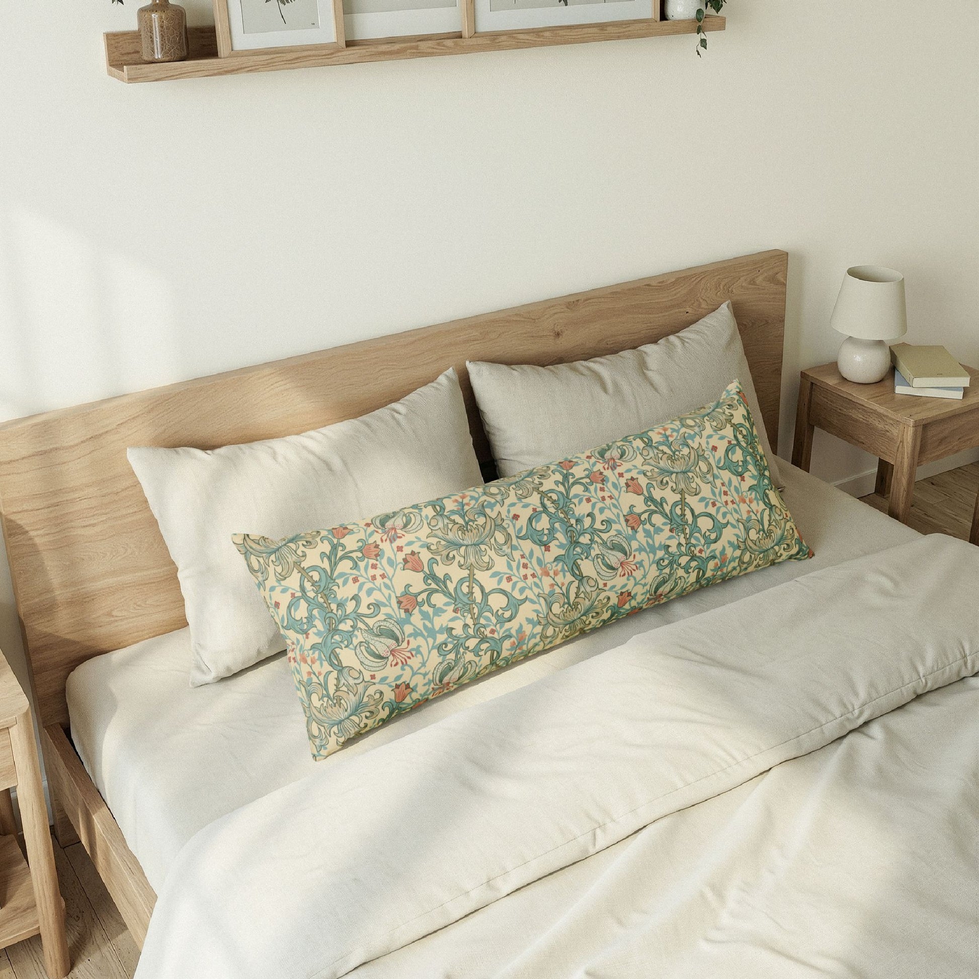 A long decorative lumbar pillow with a vintage floral and vine pattern in muted green, blue, and beige, styled on a bed with neutral bedding and a wooden headboard.