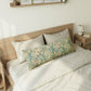 A long decorative lumbar pillow with a vintage floral pattern in green, beige, and orange, styled on a bed with white bedding and a light wood headboard.