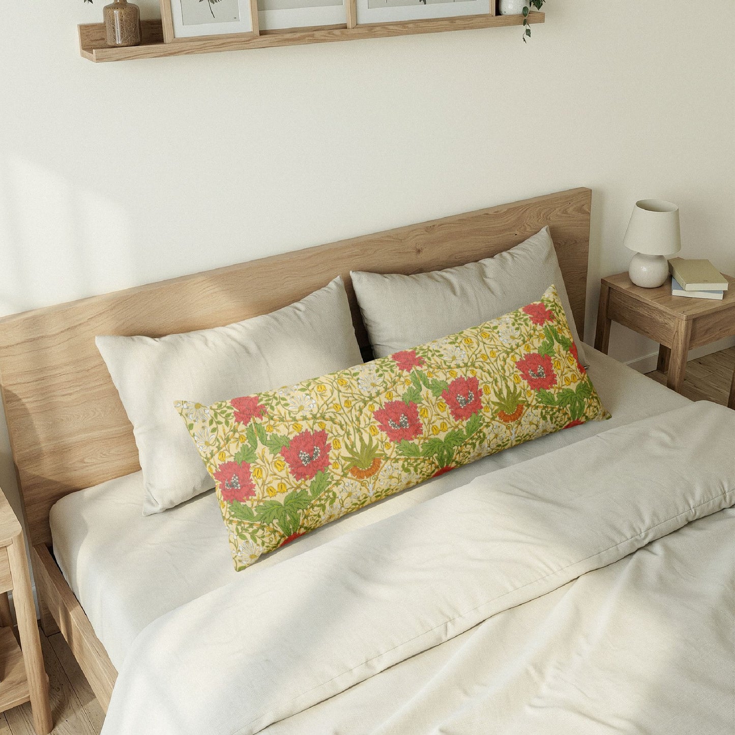 A long rectangular lumbar pillow with a colorful floral print in red, pink, yellow, and green, styled on a bed with white bedding and neutral-toned wooden furniture.
