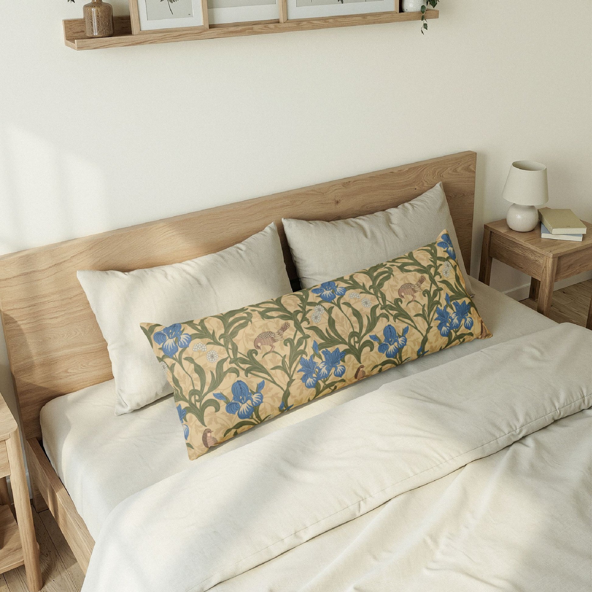 A long decorative lumbar pillow with a floral pattern in blue, green, and beige, styled on a bed with white bedding and a light wood headboard.