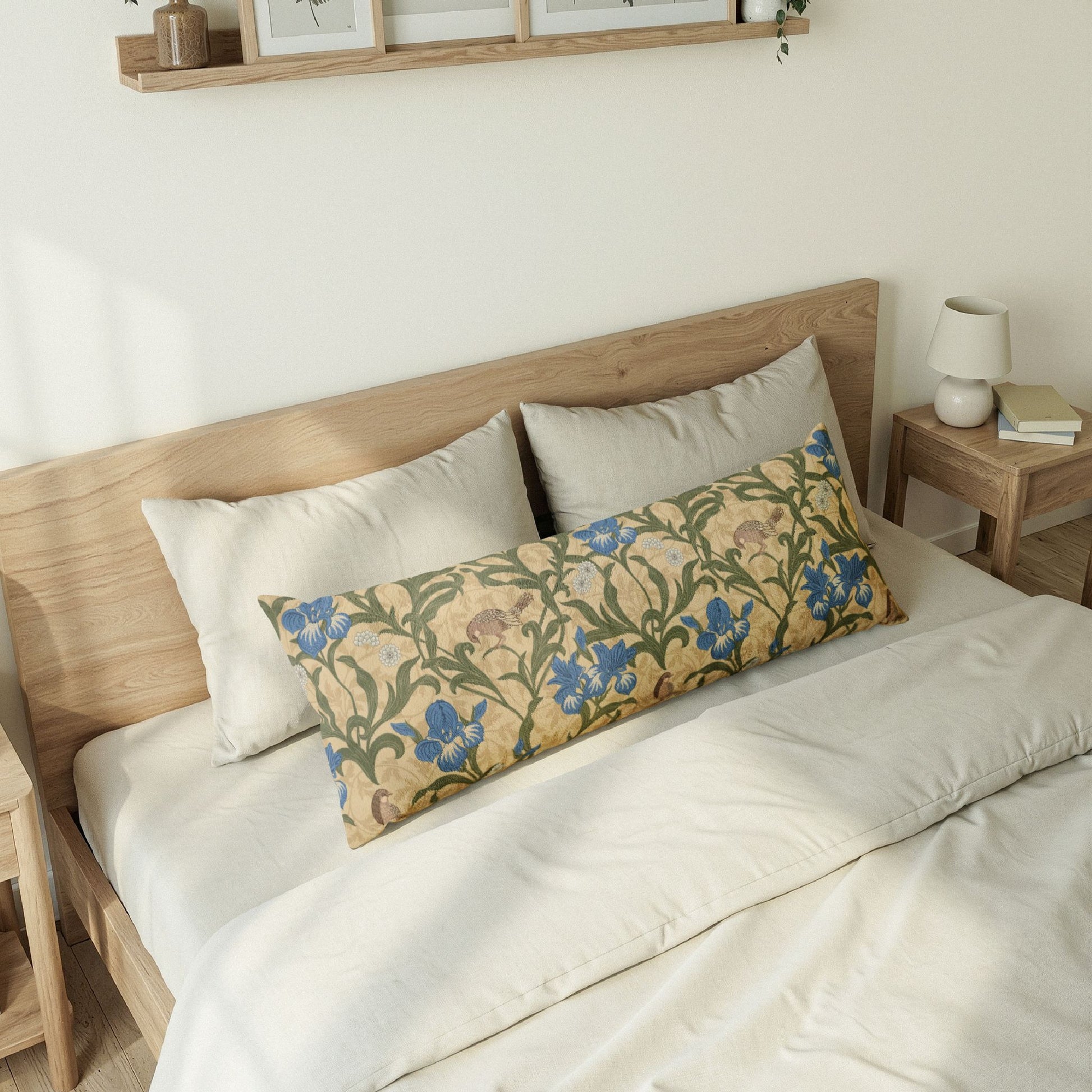 A long decorative lumbar pillow with a William Morris-style floral pattern in blue, green, and beige, styled on a bed with white bedding and a light wood headboard.