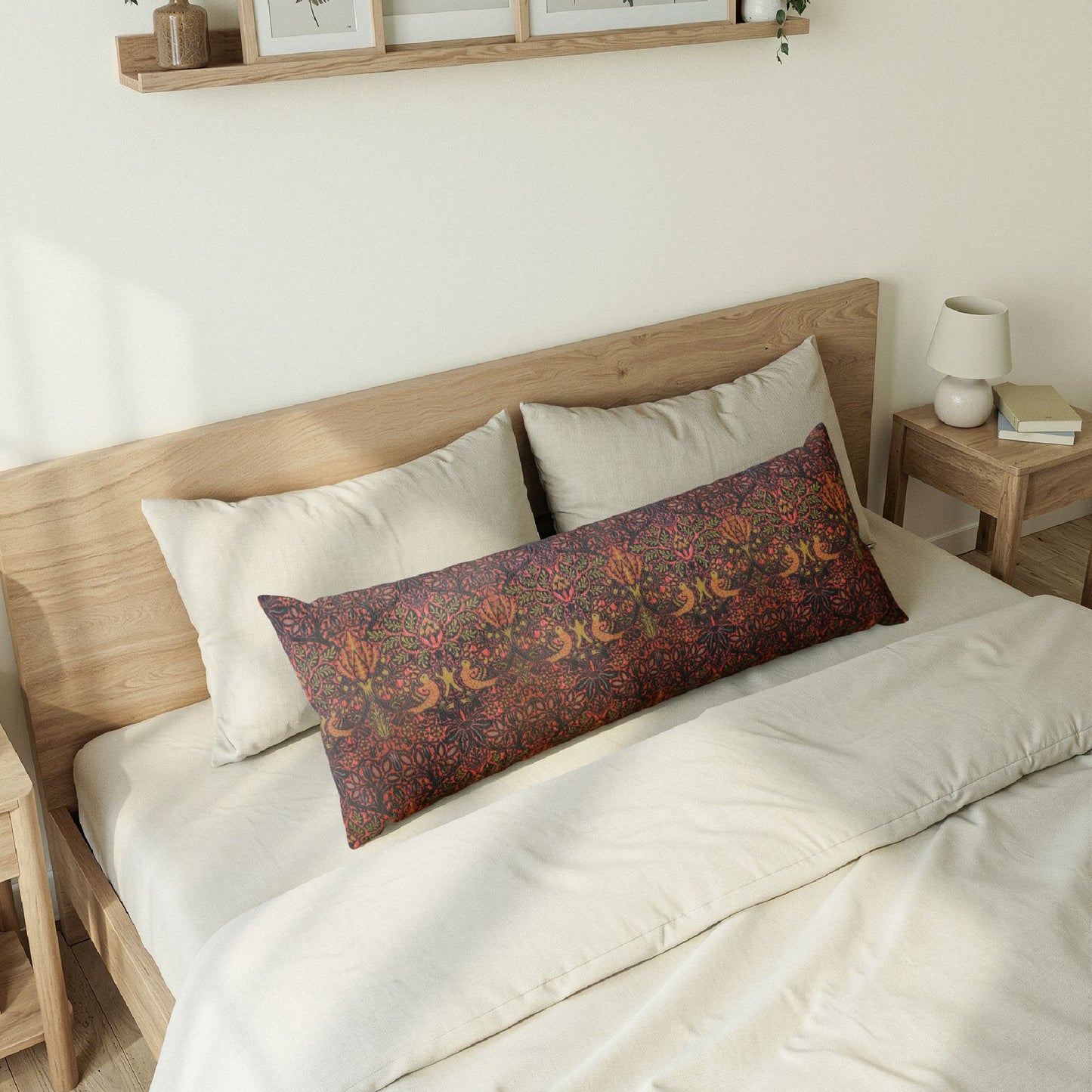A rectangular lumbar pillow with a detailed floral and bird pattern in rust, orange, and purple, styled on a bed with cream bedding and a light wood headboard in a bright, modern bedroom.