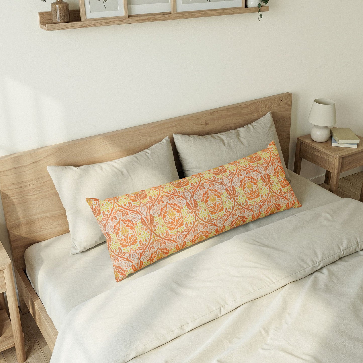 A long rectangular lumbar pillow with a warm orange and yellow botanical damask pattern rests on a bed with cream bedding and light wood headboard in a softly lit bedroom.