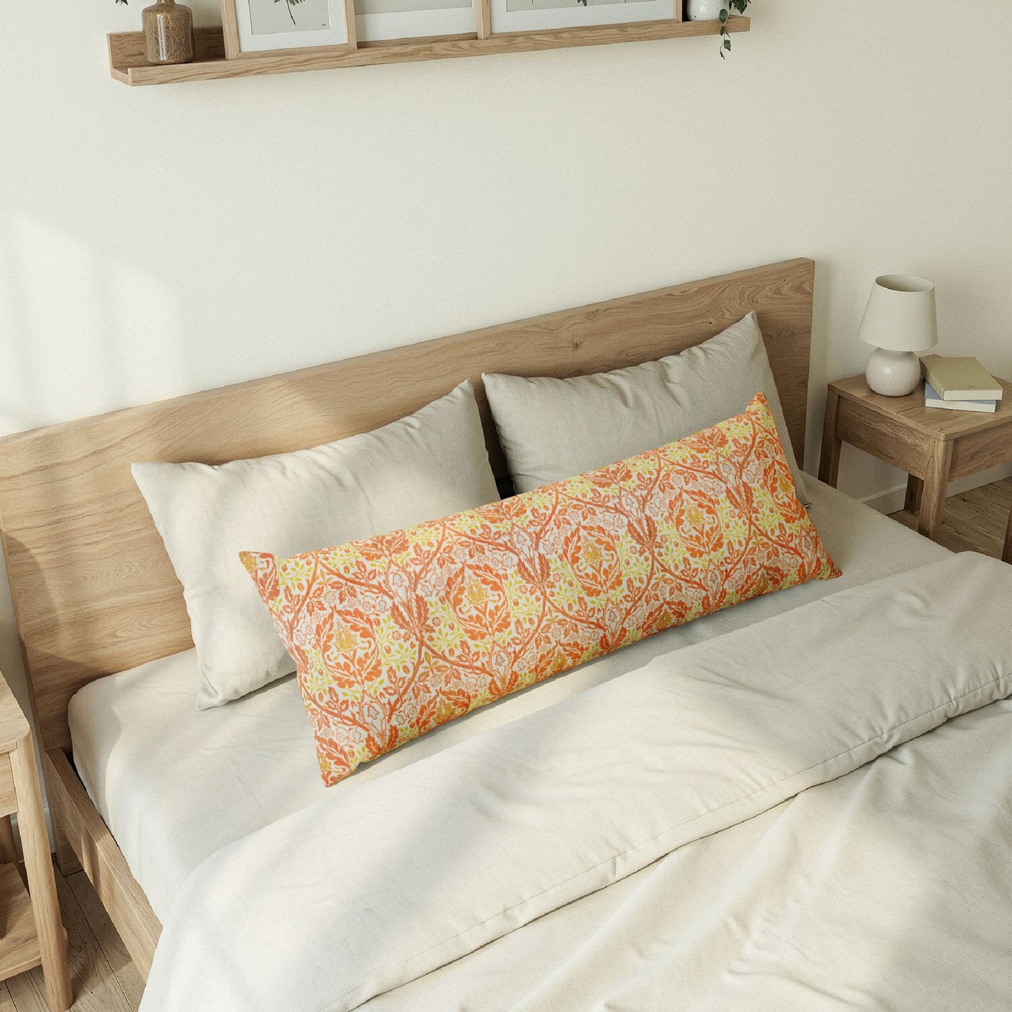 A long decorative lumbar pillow with an orange and yellow floral damask pattern rests on a bed with white bedding and light wood headboard in a softly lit bedroom.