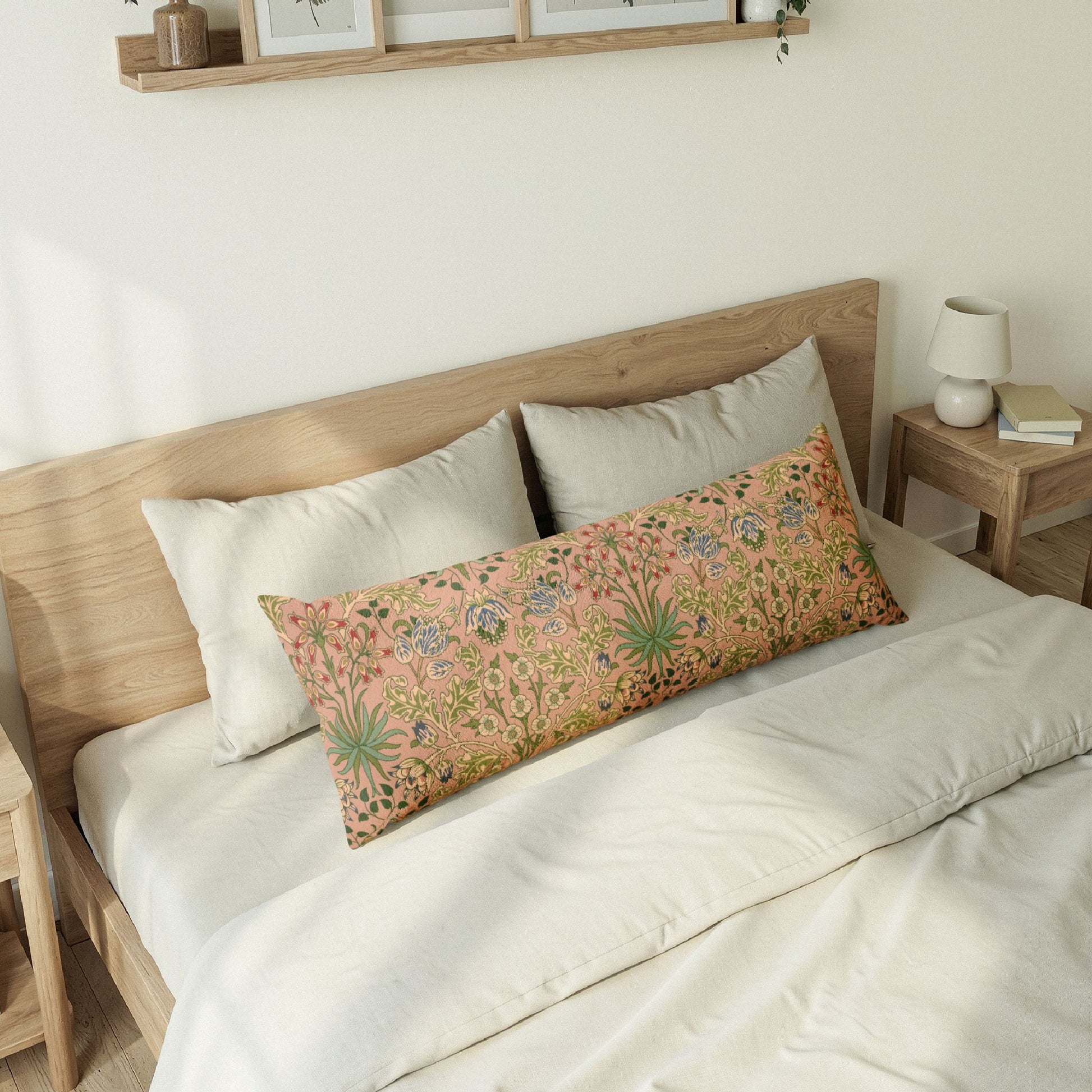 A long rectangular decorative pillow with a dense botanical and floral pattern in muted pink, green, and blue tones, styled on a bed with white linens and a light wood headboard in a softly lit bedroom.