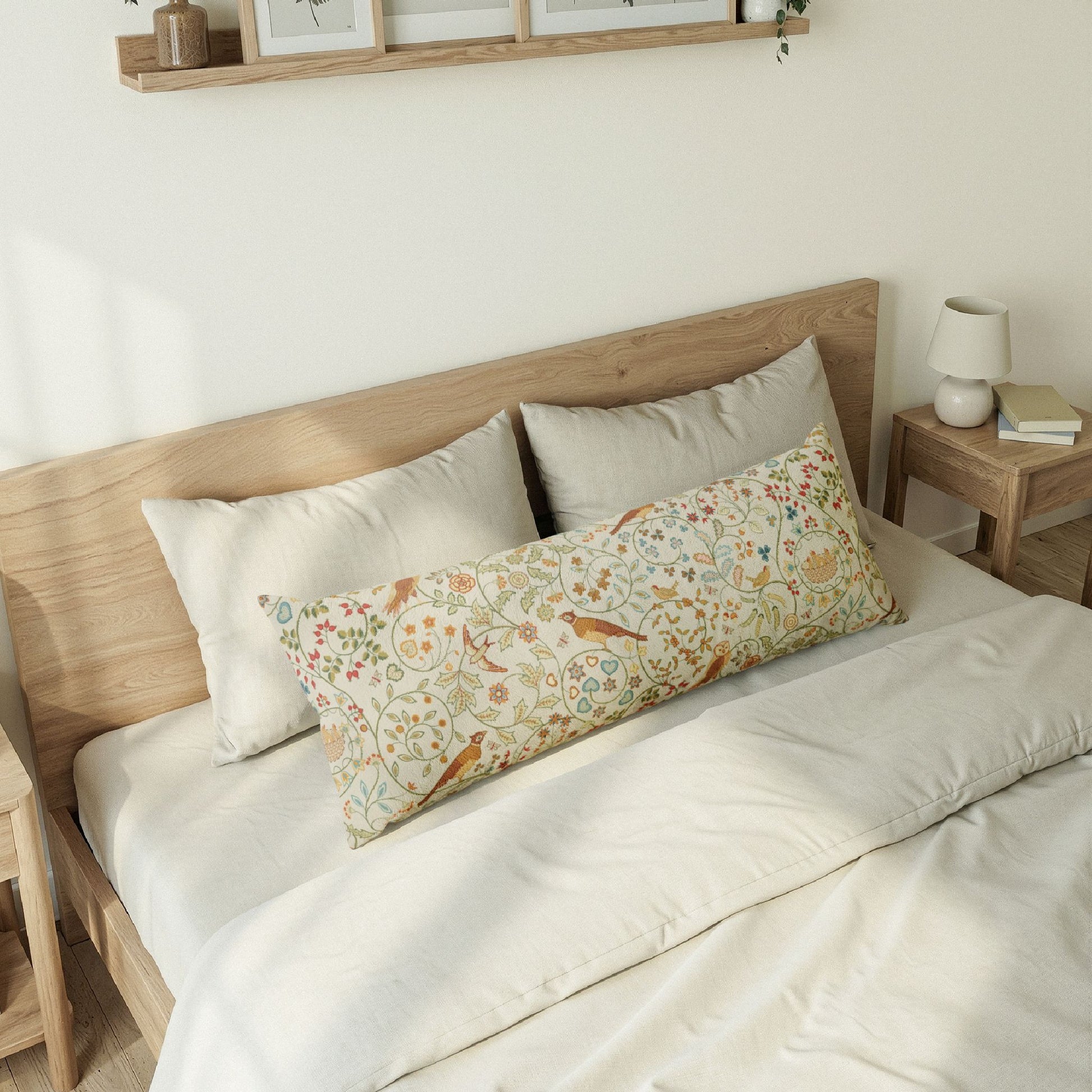 A long decorative lumbar pillow with a colorful floral and bird pattern rests on a bed with neutral cream bedding and light wood headboard in a softly lit bedroom.
