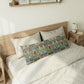 A decorative rectangular lumbar pillow with a detailed botanical pattern in blue, green, and brown, styled on a bed with cream bedding and a light wood headboard.