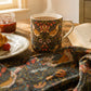 A cozy breakfast scene with a floral ceramic mug filled with steaming coffee on a wooden table, alongside a croissant, jam, butter, and an open book. A matching patterned textile is draped in the foreground, and a cabinet with teapots is visible in the background near a window.