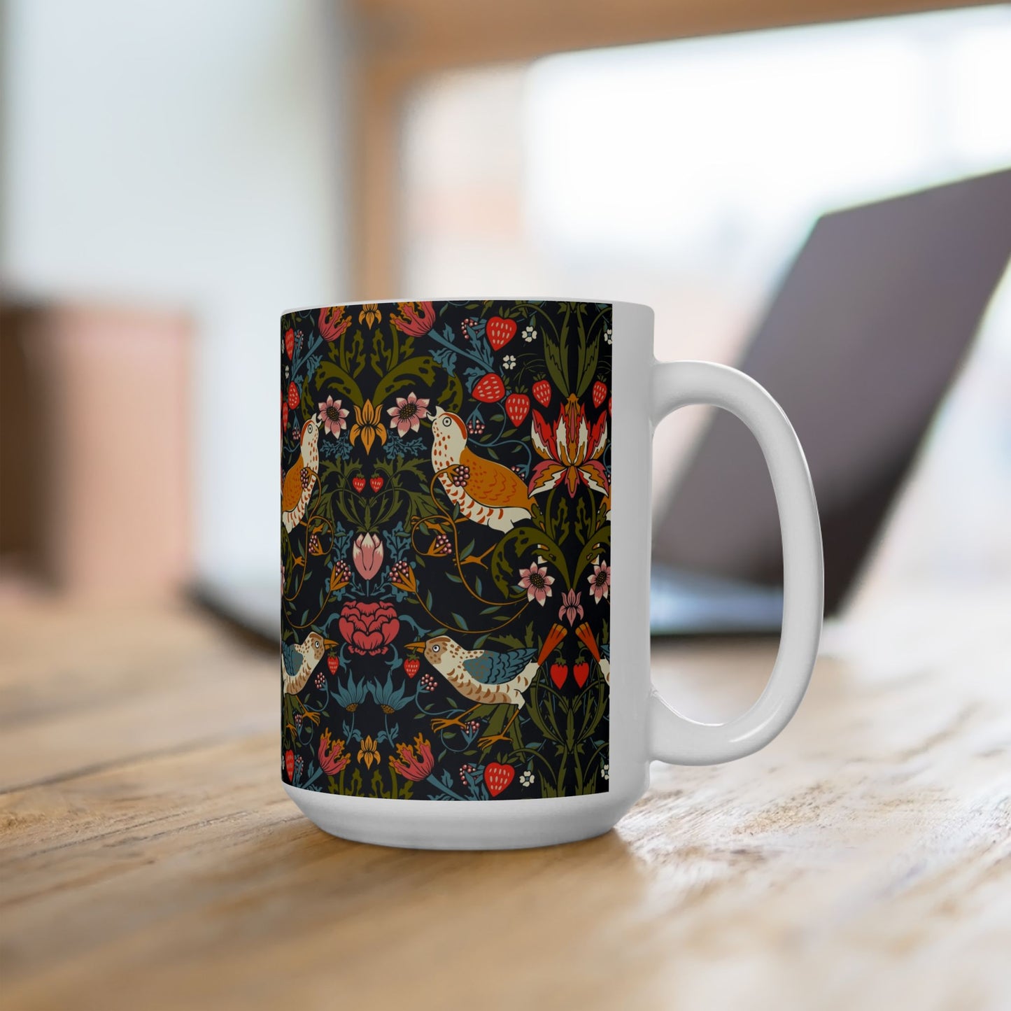 A white ceramic mug with a dark, intricate botanical and bird pattern, sitting on a wooden table with a blurred laptop in the background.